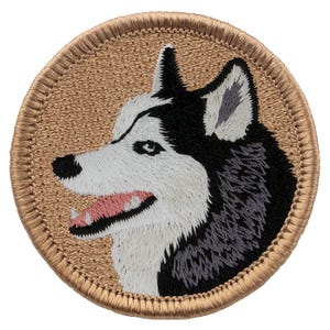 May include: A circular embroidered patch featuring a profile view of a husky dog with black, white, and grey fur. The dog has an open mouth showing a pink tongue and white teeth. The background of the patch is tan with a matching tan stitched border.