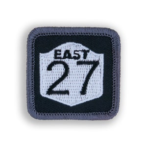 May include: A black and white embroidered patch with a gray border. The patch features a white shield with the text "EAST 27" in black.