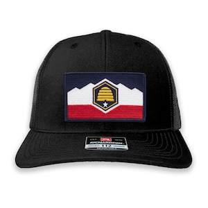 May include: Black trucker hat with a patch featuring a beehive and mountains in red, white, and blue. The patch is a stylized version of the flag of Salt Lake City, Utah. The hat is adjustable and has the Richardson 112 logo.