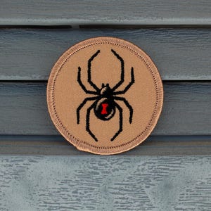 May include: A brown embroidered patch with a black widow spider design. The spider has a red hourglass marking on its abdomen.