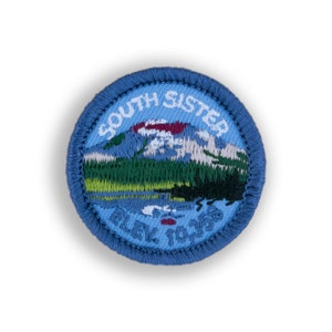 May include: A blue embroidered patch with a white outline. The patch features a mountain scene with the text "South Sister Elev. 10,358".