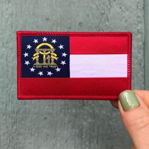 May include: A rectangular patch featuring the flag of Georgia, USA. The design includes a blue canton with stars and a gold emblem, a red field, and a white stripe. The patch is bordered with red stitching and the text "IN GOD WE TRUST".