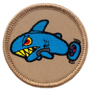 May include: Embroidered patch featuring a cartoon shark in shades of blue, with a yellow-toothed grin and a red and yellow eye. The shark is set against a tan background with a stitched border, suitable for jackets, bags, or other fabric items.