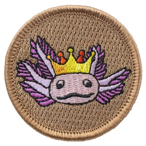 May include: A round embroidered patch with a brown background featuring a pink axolotl wearing a yellow crown with orange and red accents. The axolotl has purple gills and a smiling face.