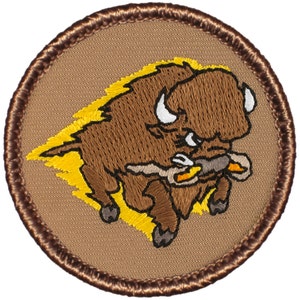 May include: A brown embroidered patch with a brown bison charging forward with a yellow flame behind it. The patch has a brown outer border.