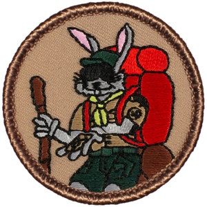 May include: A brown embroidered patch with a cartoon rabbit wearing a green hat, a red backpack, and a brown hiking stick. The rabbit is wearing a scout uniform with a yellow neckerchief.