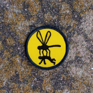May include: A yellow and black embroidered patch with a silhouette of a mosquito. The mosquito is depicted as a large insect with long legs and wings.