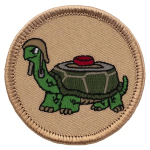 May include: Embroidered patch featuring a cartoon turtle with a green shell and body, wearing a hat and carrying a shovel. The turtle has a red object on its back. The patch is circular with a tan background and a tan border.