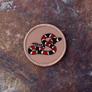 May include: A tan embroidered patch with a red, black, and yellow snake design. The snake is curled up in a circle.