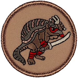 May include: A brown and gray embroidered patch of an armadillo wearing a red bandana and holding a sword. The armadillo is standing on a tan background.