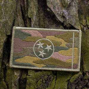 May include: A camouflage fabric patch with the state of Tennessee's flag embroidered on it. The flag features three white stars in a circle on a blue background.