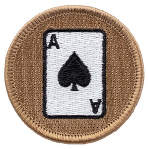 May include: A brown embroidered patch with a white playing card featuring the Ace of Spades. The card has a black spade and the letter A in each corner.