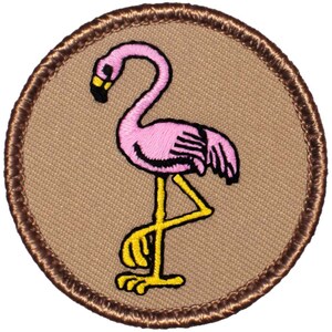 May include: A brown embroidered patch with a pink flamingo standing on one leg. The flamingo has a yellow beak and legs.