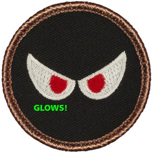 May include: A black embroidered patch with two white eyes with red pupils. The patch has a brown border and the text "GLOWS!" in green.