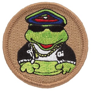 May include: Embroidered patch featuring a green frog wearing sunglasses, a gold chain, and a black jacket. The frog is wearing a blue and red hat with a white star on top. The patch is circular and has a brown background.