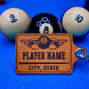 May include: A brown leather keychain with a black and white billiard ball design. The keychain features a black 8-ball, a white 2-ball, and a white 10-ball. The keychain also has a black and white image of a pool cue and the text "PLAYER NAME CITY, STATE".
