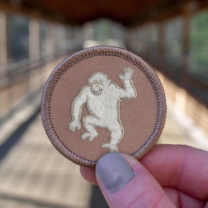 May include: A brown embroidered patch with a white outline of an ape with one arm raised.