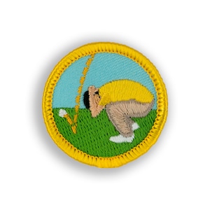 May include: A yellow and green embroidered patch with a cartoon of a person in a yellow shirt and brown pants swinging a golf club. The person is looking down at the ground and the golf club is in the air. The patch is surrounded by a yellow border.