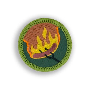 May include: A green and brown embroidered patch with a yellow and orange flame and a brown hot dog on a stick. The patch is surrounded by a green stitched border.
