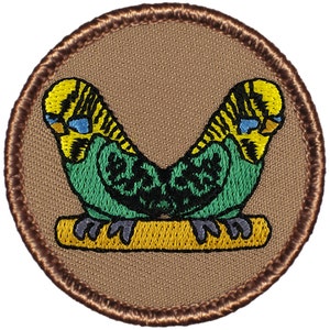 May include: Embroidered patch featuring two green and yellow parakeets perched on a branch. The parakeets are facing each other and have black eyes and blue markings on their beaks.