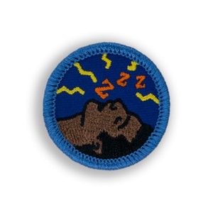 May include: A blue embroidered patch with a brown and black silhouette of a person sleeping with the word "zzz" above them. The background is blue with yellow lightning bolts.