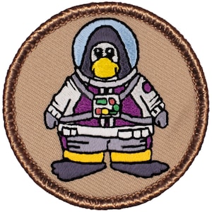 May include: Embroidered patch featuring a cartoon penguin wearing a purple and white space suit with a helmet. The penguin has a yellow beak and black eyes. The patch is brown with a white background.
