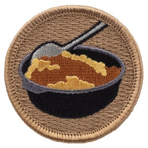 May include: A brown embroidered patch with a black bowl filled with orange and yellow food. A silver spoon is in the bowl.