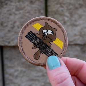 May include: A brown and yellow embroidered patch with a cartoon beaver wearing a yellow construction helmet and holding a log. The beaver is crossing a yellow caution tape.