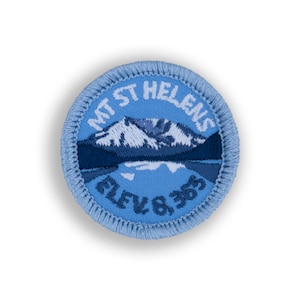 May include: A blue embroidered patch with a white outline. The patch features a mountain range with a lake in the foreground. The text "MT ST HELENS ELEV 8,365" is embroidered in white around the image.
