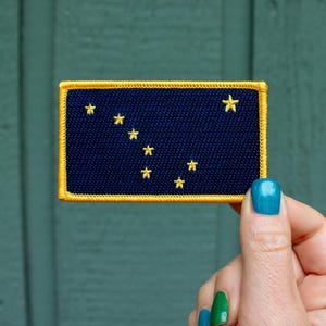 May include: Embroidered patch featuring the flag of Alaska. The patch has a navy blue background with gold stars and a gold border. The Big Dipper and the North Star are represented in the design. The patch is rectangular in shape.