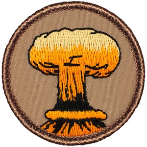 May include: A brown embroidered patch with a yellow and orange mushroom cloud design. The patch is circular and has a brown border.