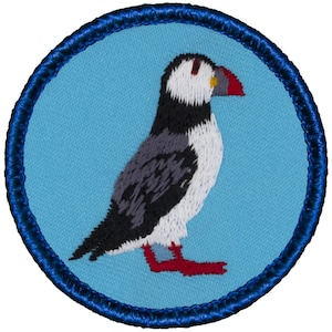 May include: A blue and white embroidered patch featuring a puffin with a red beak and red feet. The puffin is standing on a blue background and is surrounded by a blue border.