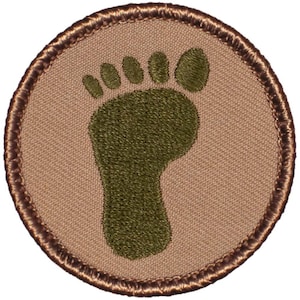 May include: A round, tan fabric patch with a brown stitched border. The patch features a green embroidered footprint.