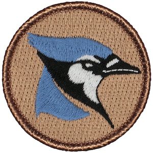 May include: A brown embroidered patch with a blue jay bird design. The bird is facing right and has a blue crest, white face, and black eyes. The patch is circular and has a brown border.