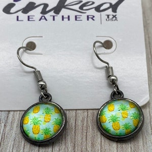 May include: A pair of silver dangle earrings with a round, blue and green glass charm featuring a pineapple pattern. The earrings are on a white card with the text "inked leather tx usa".