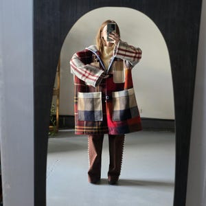 May include: A patchwork coat with a hood and large pockets. The coat features a variety of plaid patterns in red, brown, and beige. The person is wearing brown pants. The coat is reflected in a large, arched mirror.