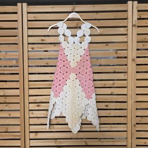 May include: A handmade dress with a unique design. The dress is made of white and pink circular fabric pieces, arranged in a geometric pattern. It hangs on a wooden hanger against a wooden slatted backdrop. The dress has a sleeveless design.