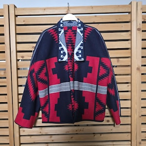 May include: A patterned jacket in red, navy blue, and gray, hanging against a wooden background. The jacket features geometric designs, including zigzags and rectangular shapes. The collar and edges are trimmed with a white and gray pattern.