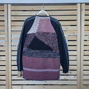 May include: A color-blocked sweater in shades of black, white, pink, and burgundy hangs on a hanger. The sweater features a geometric design with a mix of textures, including knit and crochet elements. The sleeves are solid black with a thin white stripe.