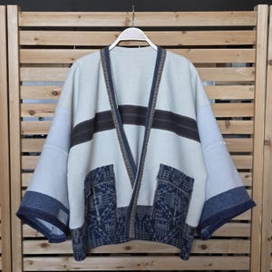 May include: A light-colored kimono-style jacket with a dark brown horizontal stripe across the chest and patterned pockets. The sleeves and trim are a contrasting blue. The jacket is hanging on a white hanger.