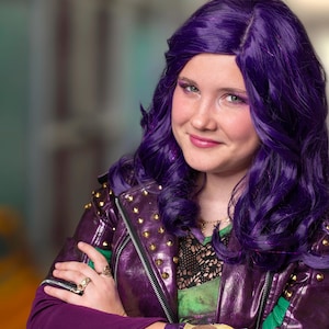 May include: A person with purple hair and makeup, wearing a purple leather jacket with gold studs, a green and black patterned top, and a purple sleeve. The person is wearing rings and a gold wrist cuff.