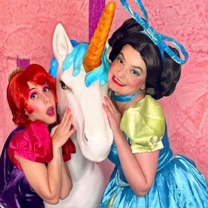May include: Two girls dressed as princesses are posing with a white unicorn statue. One girl is wearing a purple and pink dress and the other is wearing a blue and green dress.