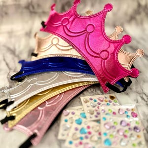 May include: A stack of colorful, metallic-looking crowns in pink, gold, blue, silver, and yellow. Each crown has a decorative design and a black elastic band. Several sheets of heart-shaped and gem stickers are in the foreground.