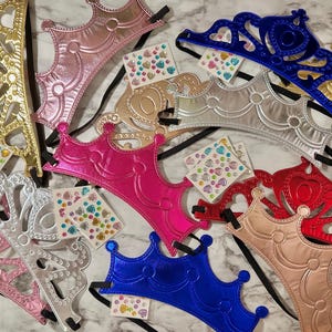 May include: An assortment of colorful toy crowns in gold, pink, silver, blue, and red. Each crown has a black elastic band and is decorated with heart-shaped embellishments. Some crowns have heart-shaped cutouts.
