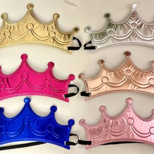 May include: Six metallic crowns in gold, silver, pink, rose gold, blue, and light pink. Each crown has a black elastic band for securing. The crowns have a classic royal design with rounded points and decorative stitching.