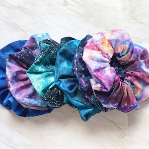 May include: A set of five galaxy print scrunchies in shades of blue, purple, and pink. The scrunchies are made of soft fabric and have a stretchy elastic band.
