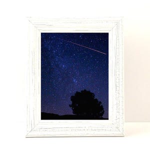 May include: White framed print featuring a night sky filled with stars and a shooting star. A dark silhouette of a tree and landscape is at the bottom. The frame has a distressed, rustic look.