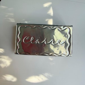 May include: A rectangular silver-toned metal tin with a wavy, scalloped border embossed around the perimeter. The center of the lid features the name Claire written in a raised, cursive script font. The surface has a reflective, metallic finish.