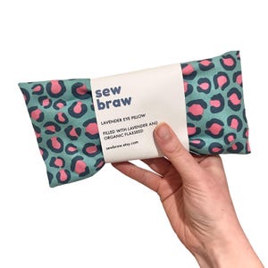 May include: A lavender eye pillow with a teal leopard print and pink spots. The pillow is held by a hand and has a white label that reads "sew braw" and "LAVENDER EYE PILLOW FILLED WITH LAVENDER AND ORGANIC FLAXSEED".
