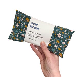 May include: A teal blue eye pillow with a white label that says "sew braw" and "Lavender Eye Pillow Filled with Lavender and Organic Flaxseed sewbraw.etsy.com". The pillow has a green, pink, and yellow abstract pattern.